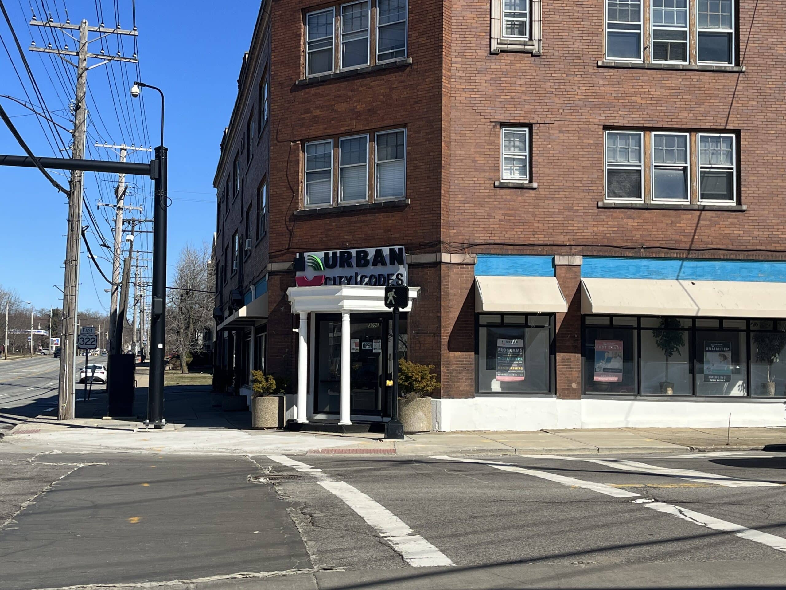 The exterior of the Urban City Codes building from a view across the street. The organization is housed in a brick building with blue and tan awnings and a sign that reads “Urban City Codes” alongside the organization’s logo. The entrance to the space has two thin white pillars leading to the doorway.