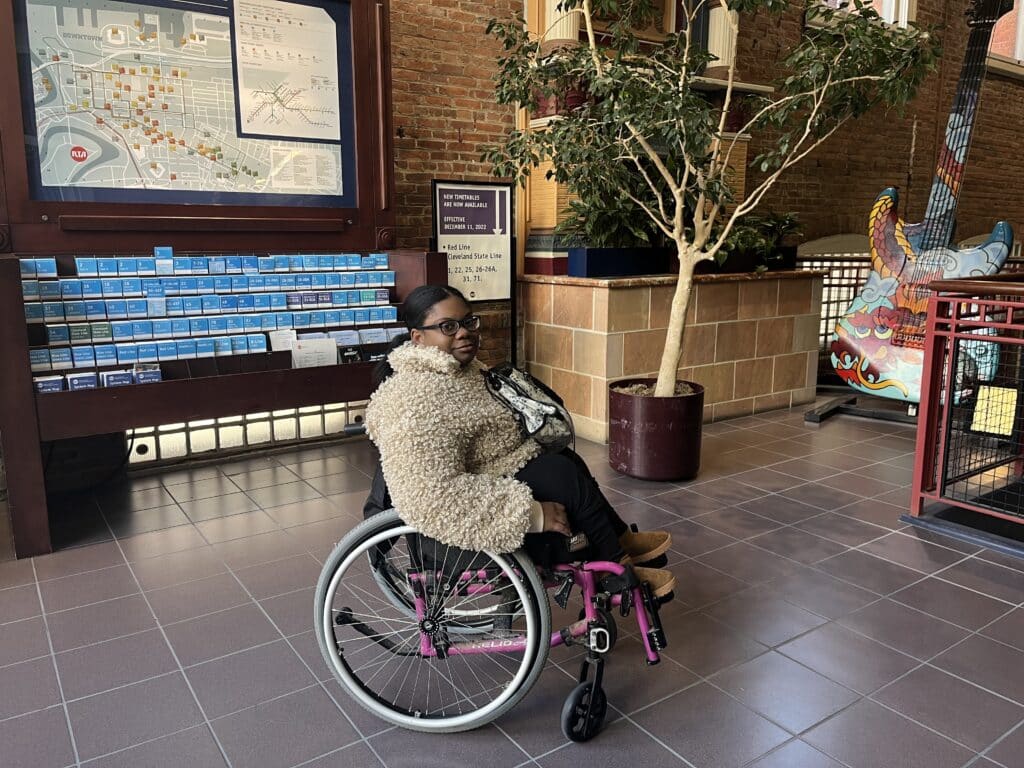 Shalida Dobbins, who uses a wheelchair, waiting in the lobby of RTA's main office near a map of RTA routes, a potted tree, and a painted guitar. 