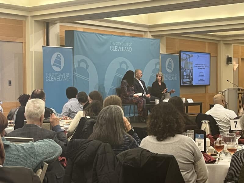 A crowd of people seated at circular tables watch as Robin Steinberg, the founder and former CEO of The Bail Project, speaks on a stage with a blue City Club backdrop, seated next to panel moderator Ayesha Bell Hardaway and the organization’s current CEO David Gaspar.
