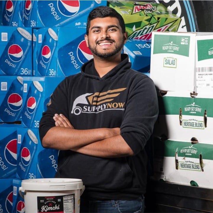 SupplyNow founder Aaron George smiling and posing with his arms folded across his chest as he stands in front of cases of Pepsi and Mountain Dew and boxes of salad and near a bucket of kimchi. 