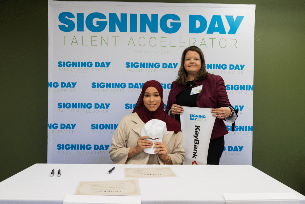 Yasmin Abdul-Khaliq sitting at a table holding a Keybank baseball cap in front of a “Signing Day” backdrop next to KeyBank’s Cyber Defense Center director, Jennifer Zacharias, who is holding a KeyBank pennant flag. 