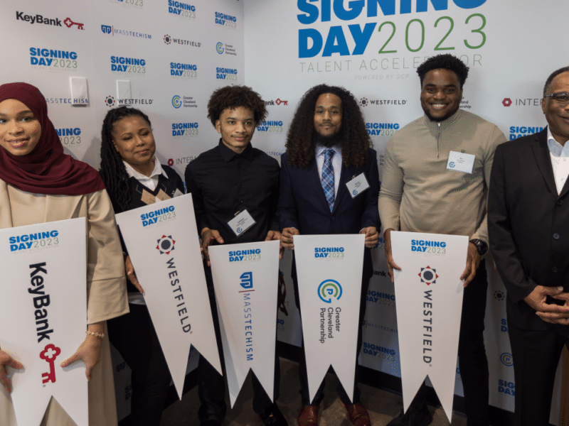 Five apprentices in the talent accelerator holding pendants that say the name of their workplaces and standing in front of a backdrop that reads “Signing Day 2023” with Craig Platt, managing director of GCP’s IT sector partnership. From left to right, the pendants read “KeyBank, Westfield, Masstechism, Greater Cleveland Partnership, and Westfield.”