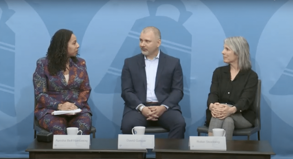 Ayesha Bell Hardaway speaking to David Gaspar and Robin Steinberg during the City Club forum. The three are sitting behind a blue City Club backdrop in front of a coffee table with three mugs and name tags.