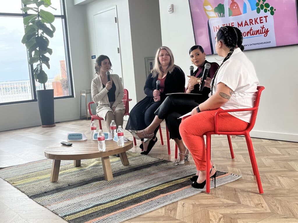Panelists sitting on red chairs and holding microphones behind a small table with bottled water and name plates. A screen in the background shows an illustration of a grocery bag filled with fresh food.