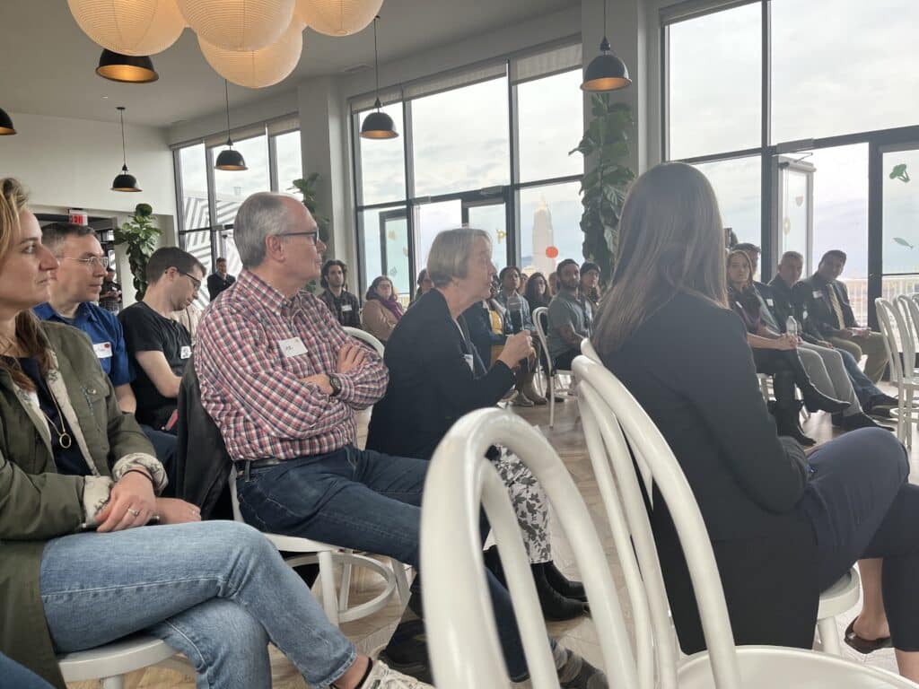 An audience member seated in the second row holds a mic while asking a question as the rest of the audience, seated in rows of white chairs, listens. 