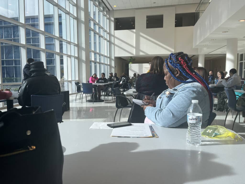 A community member takes notes during the meeting as sunlight pours in from a wall of windows. 