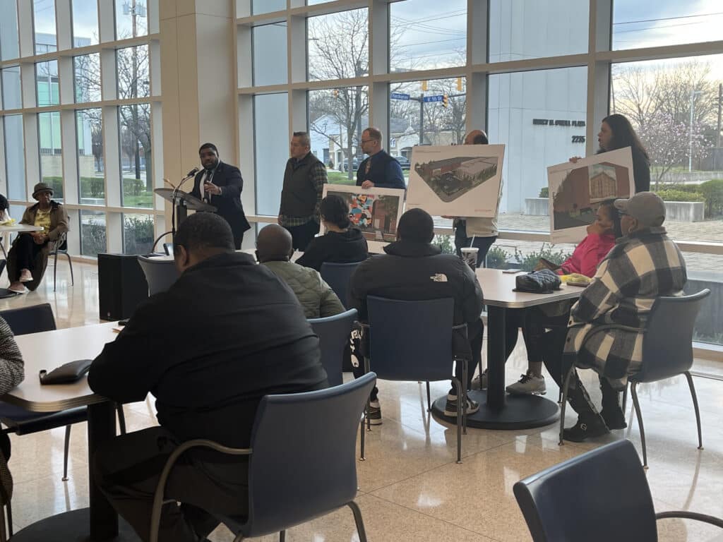Ward 5 Council Member Richard Starr talks into a microphone at a podium, standing next to leaders from International Food Solutions who are holding signs with project plans in front of residents seated at tables throughout the room. 