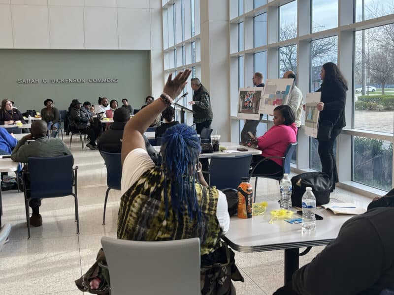 A community member raises their hand to ask a question while International Food Solutions co-president Lincoln Yee speaks at a podium and other project leaders hold up signs showing the plans.
