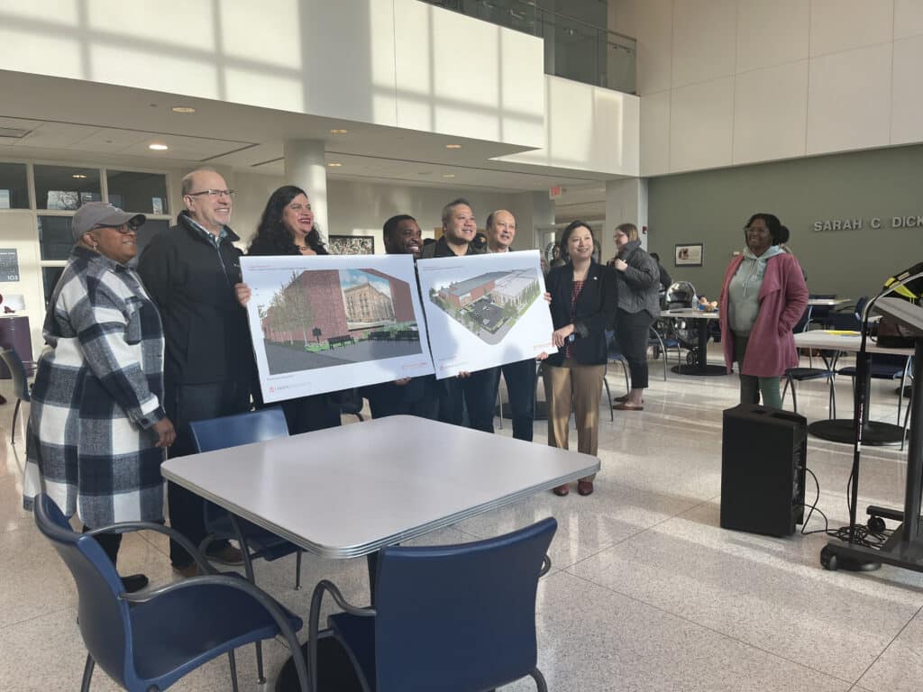 Community members and city and Project Renaissance leaders holding signs showing renderings of the chicken cooking facility and smiling for a photo as another community member watches. 