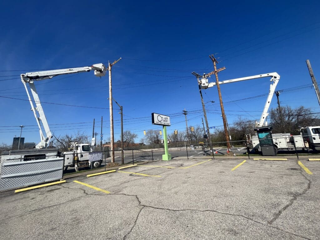 Two lineworkers working on power lines towering over a Crust pizza sign.