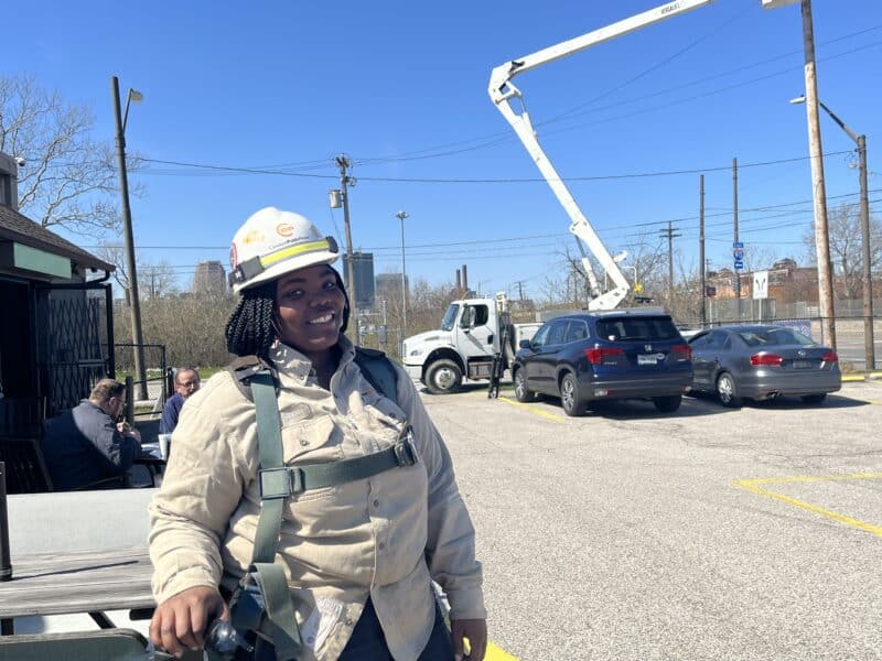 Ta’Jahnae Buchanan smiles for a photo at a job site, as other lineworkers in a bucket truck work on the power lines.