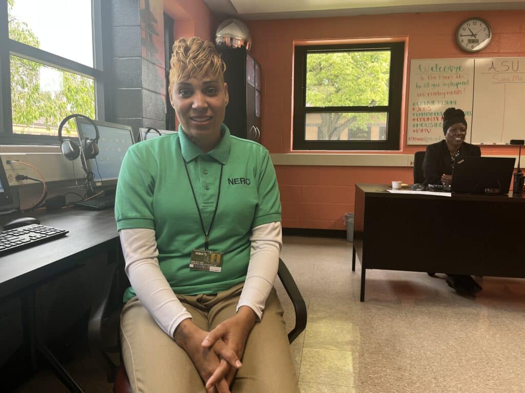 Karenda Hutsenpiller smiling sitting on a chair near the computers at the Ohio Means Jobs Center. The job center’s coordinator, Dawn Ballard-Little smiles while sitting at her desk in the background. 