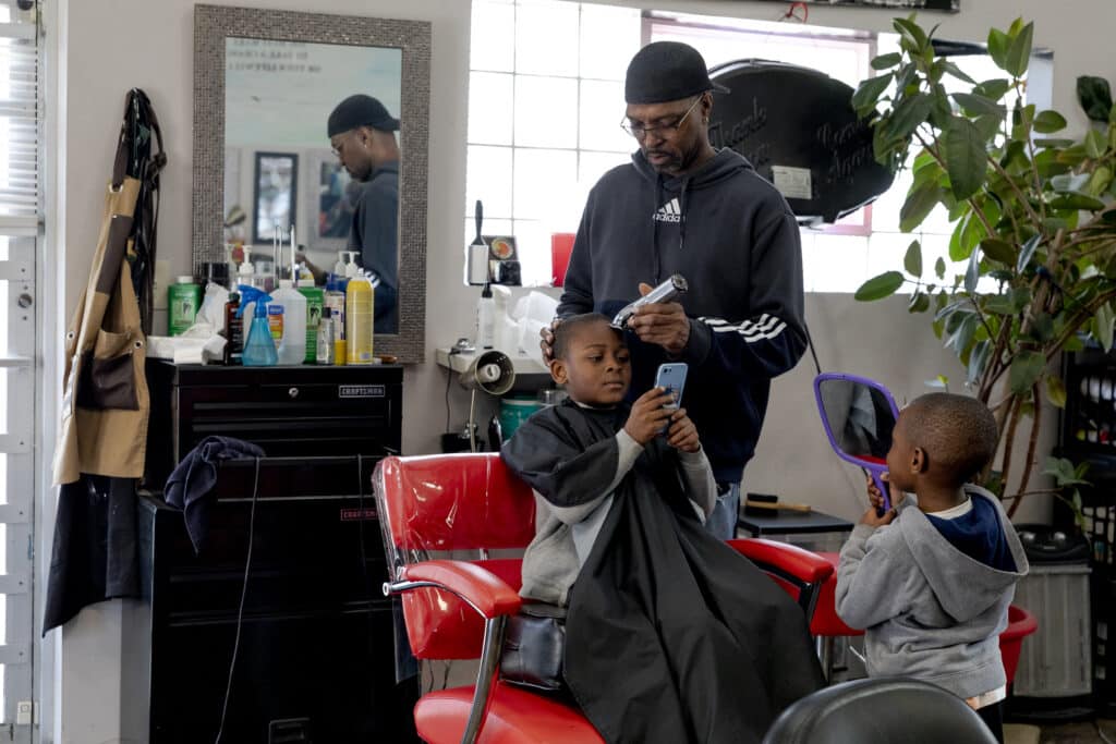 A Black man wearing a black sweater, glasses and hat, uses a clipper to cut a Black boy’s hair inside a barbershop. Another Black boy, wearing a gray sweater, stands next to the barber’s chair while holding a mirror. 