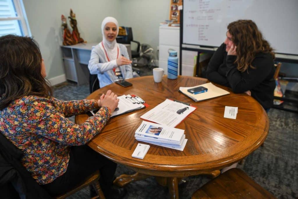 Three people sitting around a circular table near a white board. There are stacks of pamphlets and notepads on the table. 