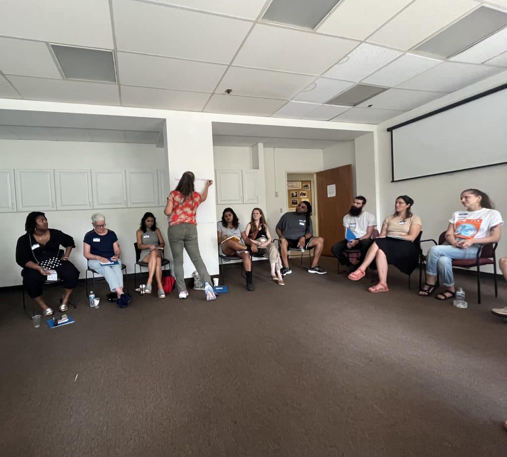 People seated in chairs in a circle as Morgan Taggart writes a discussion question on a large paper taped to the wall. 