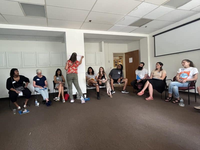 People seated in chairs in a circle as Morgan Taggart writes a discussion question on a large paper taped to the wall.