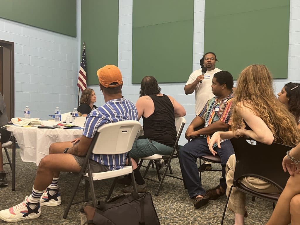 A Cooperation Jackson member standing and talking into a microphone as audience members listen. 
