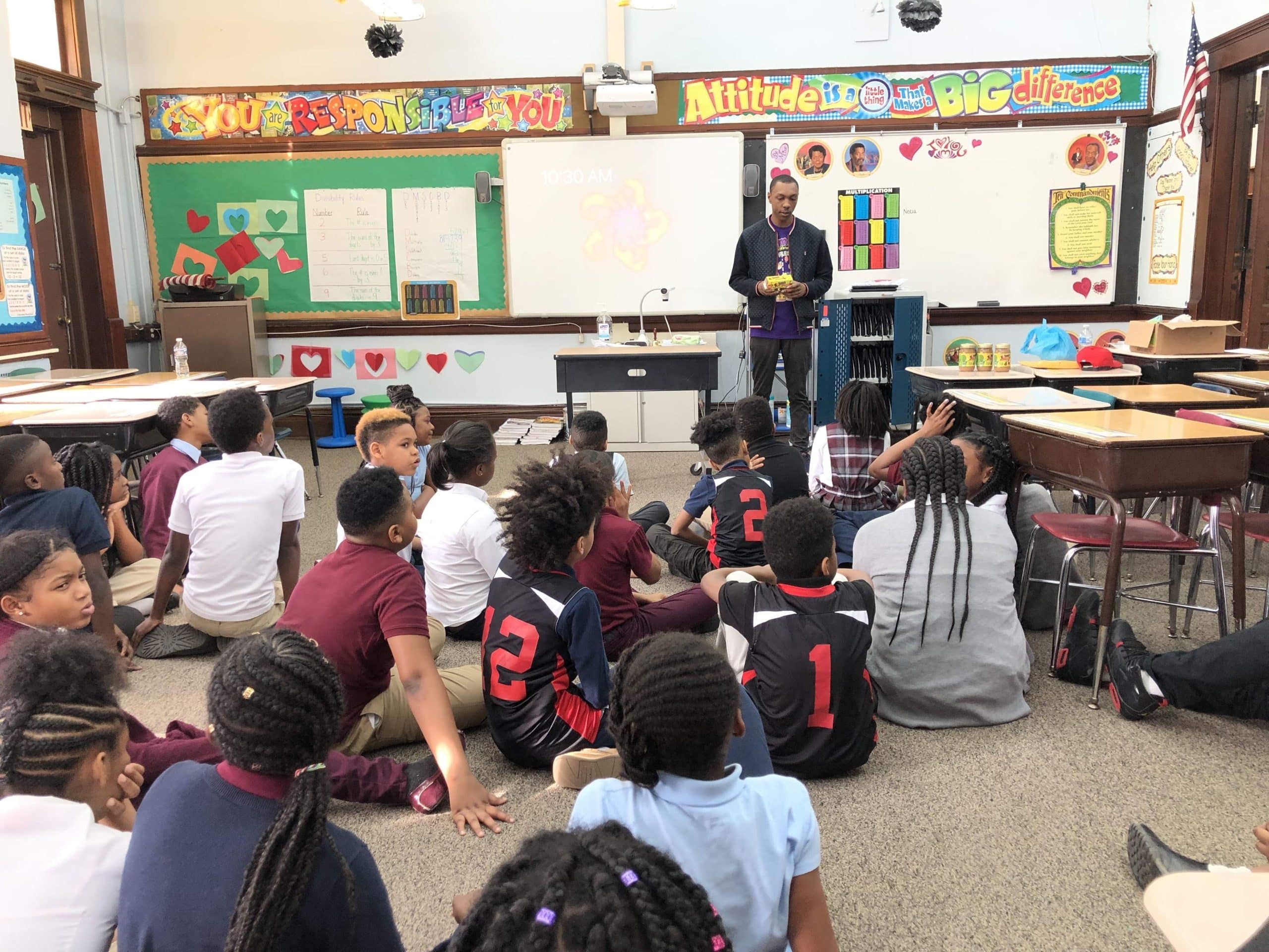 Students sitting on the floor in a classroom as Ethan Holmes stands at the front of the room giving a presentation and holding a package of applesauce.