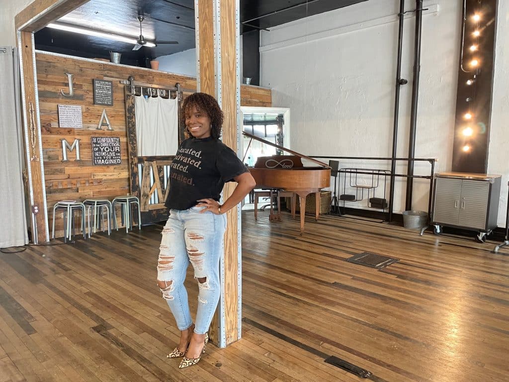 Imaj'n'Nation MultiPod owner Jami Taylor leans against a beam in her studio. She is wearing a black t-shirt and ripped jeans.