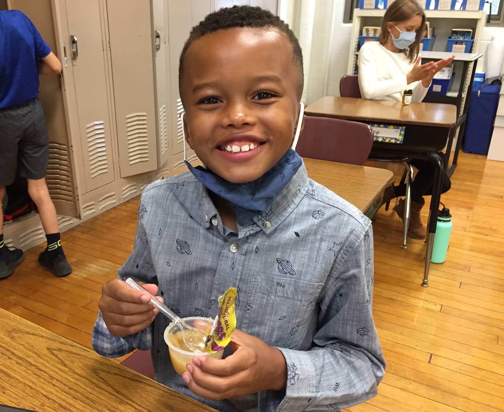 A child sitting at a desk, with a mask hanging from his chin, smiling while holding a cup of applesauce and a spoon.