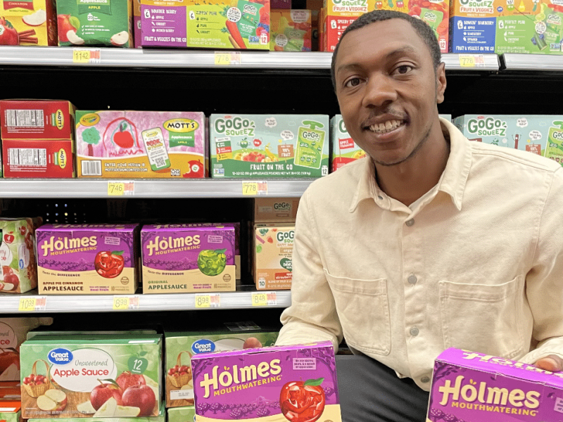 Ethan Holmes crouching down and holding two packages of Holmes Mouthwatering applesauce in a store, among shelves of applesauce.