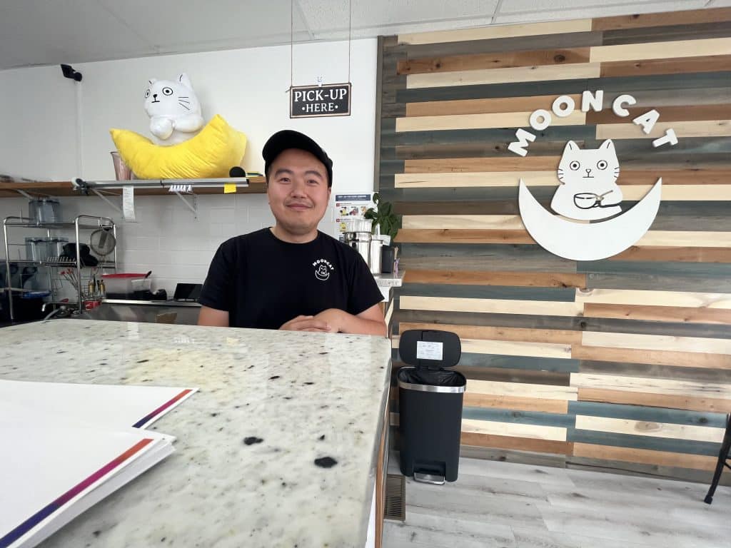 Owner Chun Wong at the counter at Mooncat Boba Cafe in between a stuffed animal version of Mooncat’s logo and a logo hanging from the wall.