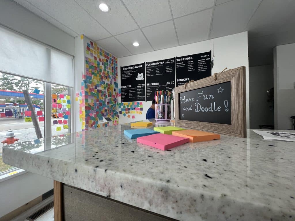 A sign that reads, “Have Fun and Doodle!” next to stacks of Post-it notes on the counter, with the Post-it note wall and menu in the background. 