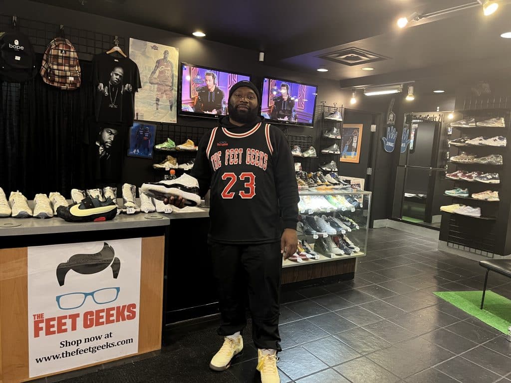 A Black man holds an athletic sneaker in front of a checkout counter. He is standing in a store with shelves and glass cases full of sneakers.