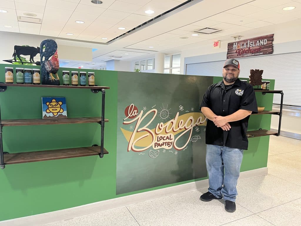 Hola Island Co. owner Luis Roman standing in front of a wall that reads, "La Bodega: Local Pantry" and a shelf with jars of Caribbean marinades.