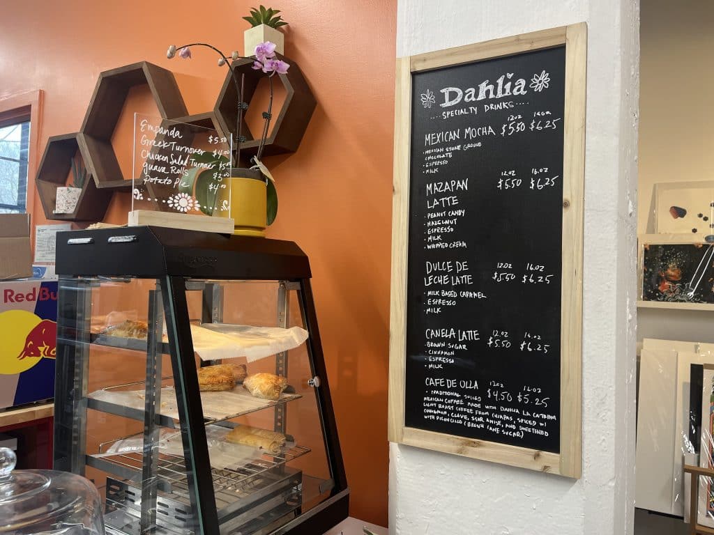The pastry case and menu board at the coffee shop. The pastries include empanadas ($5.50), Greek turnovers ($4), chicken salad turnovers ($5), guava rolls ($3), and potato pies ($4), and the specialty drinks include the Mexican Mocha, Mazapán Latte, Dulce de Leche Latte, Canela Latte, and Café de Olla. The lattes and mocha cost $5.50 for 12 oz and $6.25 for 16 oz, and the Café de Olla costs $4.50 for 12 oz and $5.25 for 16 oz.