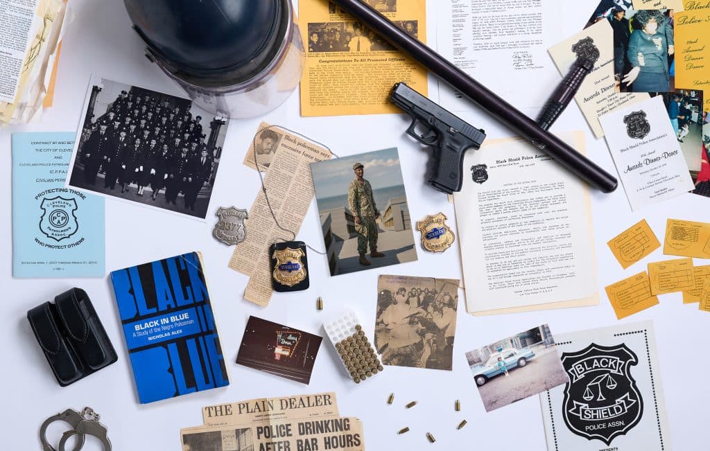A collage of items related to policing, the Black Shield Police Association and the author’s family. The items include: a police helmet; a baton; a gun; bullets; a sergeant’s badge; a newspaper clipping related to police misconduct and another related to use of force by police; and a photo of former police officer Vincent Montague.