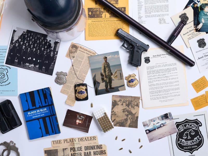 A collage of items related to policing, the Black Shield Police Association and the author’s family. The items include: a police helmet; a baton; a gun; bullets; a sergeant’s badge; a newspaper clipping related to police misconduct and another related to use of force by police; and a photo of former police officer Vincent Montague.
