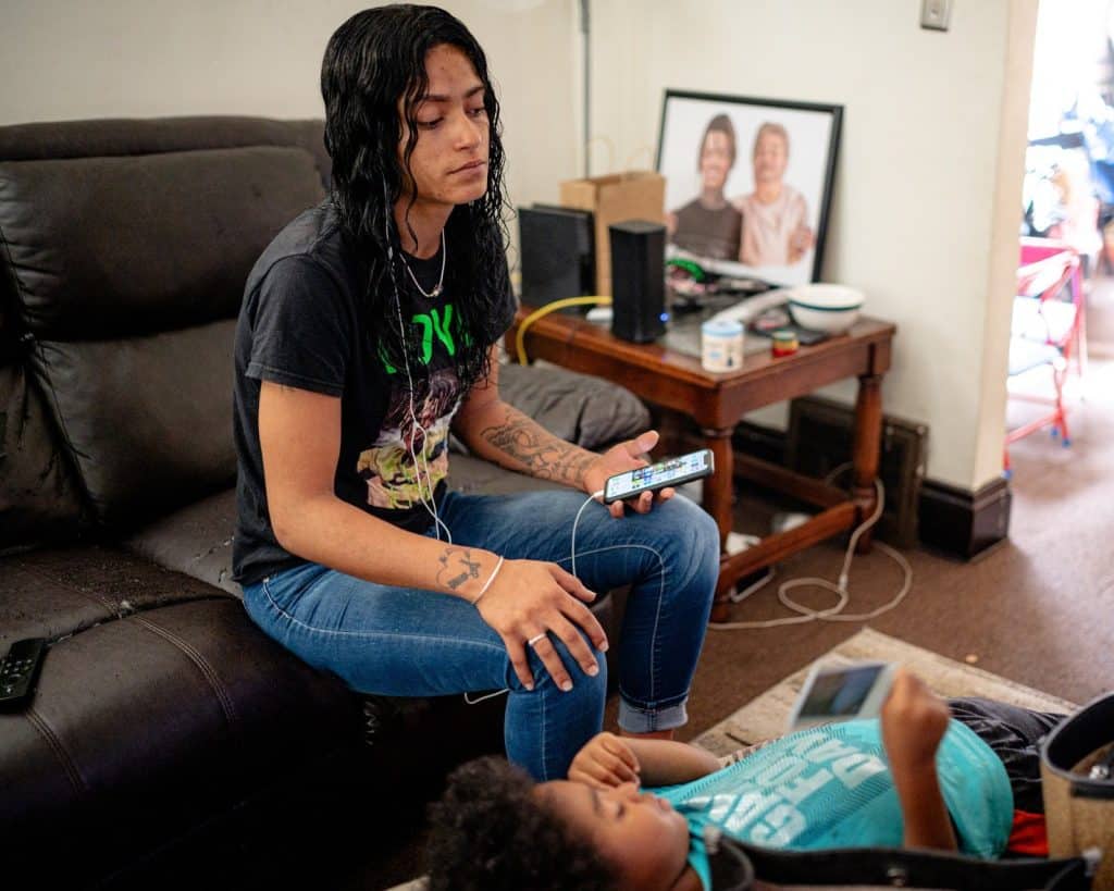 A photo of a young Black woman sitting on a couch while listening to audio from her phone through wired headphones. A Black boy with a turquoise shirt is laying on the table in front of her. 
