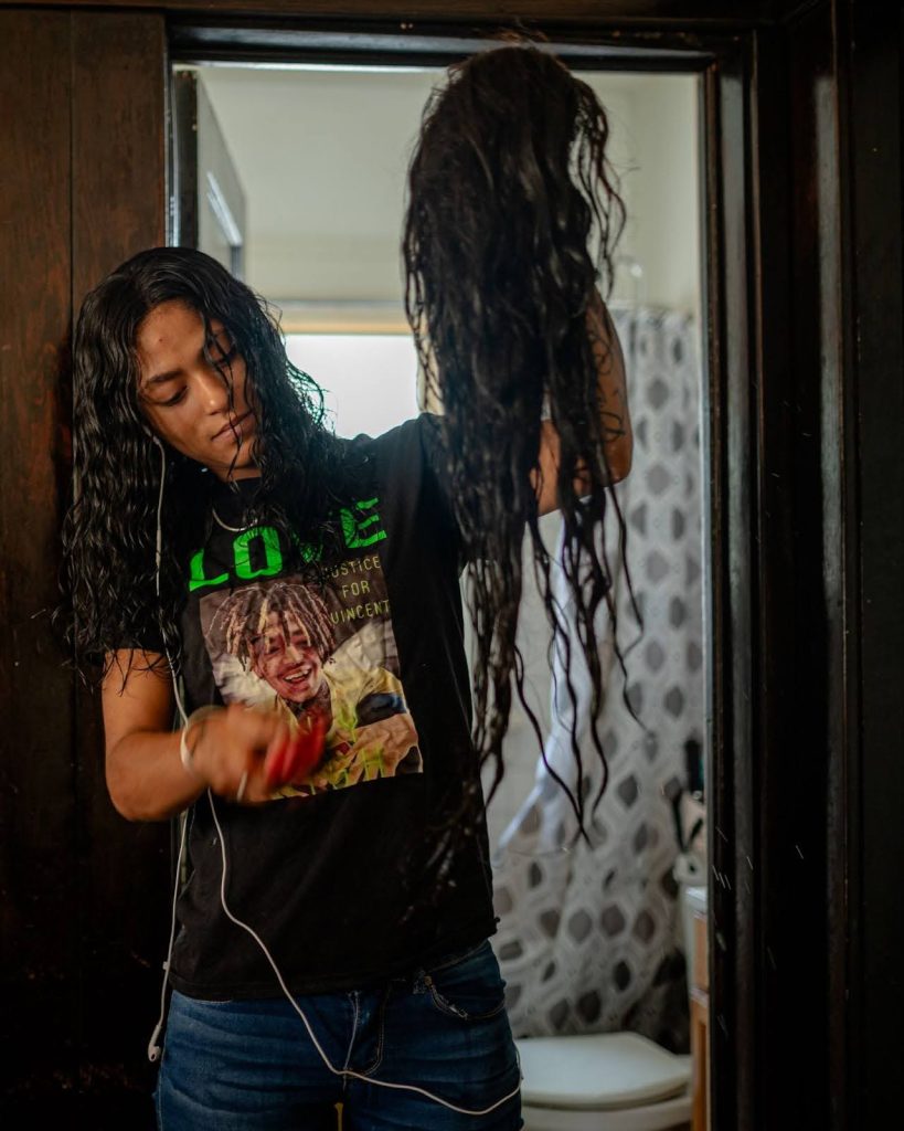 Left: A photo of a young Black woman, wearing a shirt with a photo of a Black man. The woman is holding up and brushing a wig.
