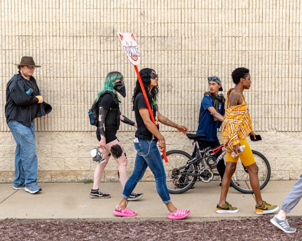 A photo of a young Black woman with sunglasses walking during a protest while holding a sign with a picture of a pig and the word “PUNKS.” 