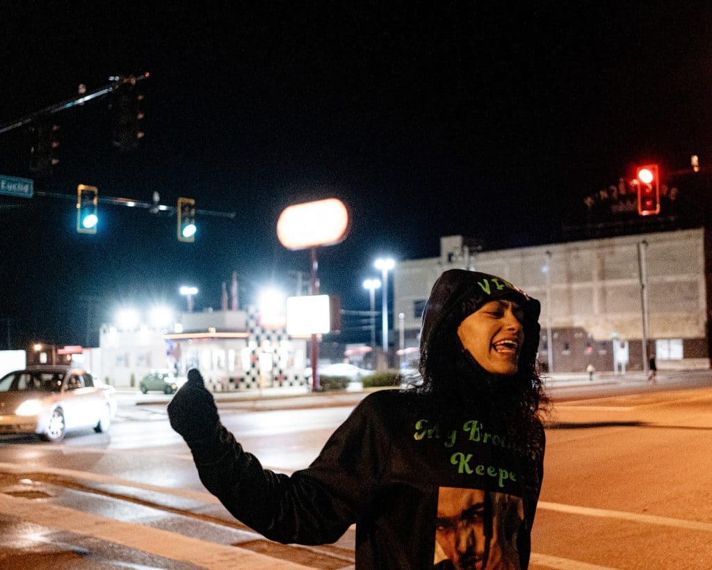 A photo of a Black woman shouting with her fist up while wearing a black hoodie with green text and an image. The woman is standing near an intersection with streetlights at night.