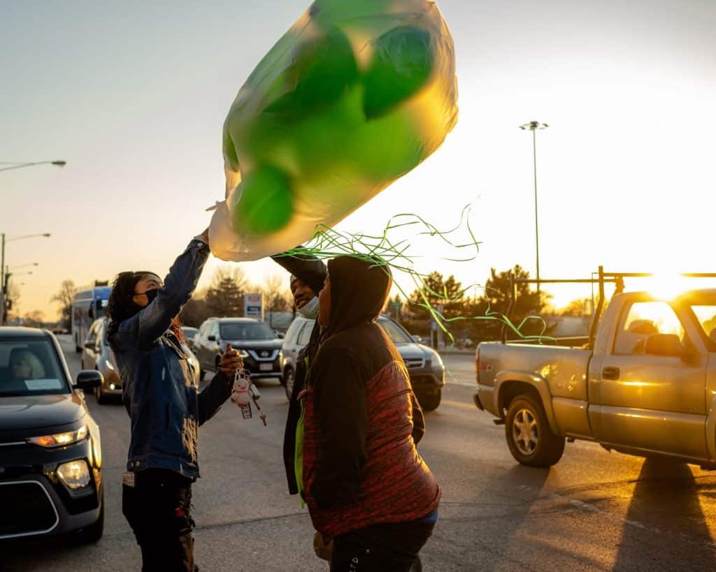 A photo of three people standing in the middle of a street filled with cars, holding a bag of green balloons as the sun is setting.