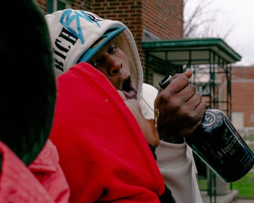 Right: A photo of a young Black man wearing a white hoodie and blue hat, pouring Hennessy into the mouth of another young Black man wearing a red hoodie. 
