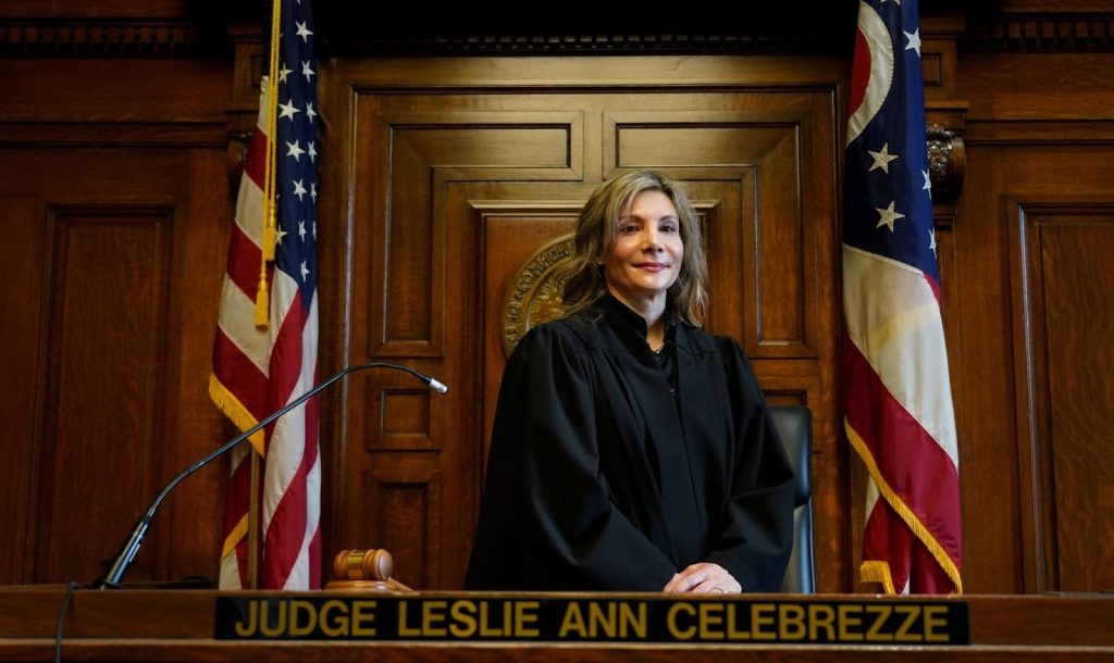 Judge Leslie Ann Celebrezze, a White woman wearing a judge’s robe, stands between two American flags. A plaque on the desk in front of her reads: “Judge Leslie Ann Celebrezze.”