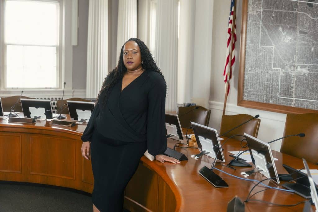 Alt text: A Black woman with long black curly hair wearing a black dress stands in front of a brown desk used by the city council in Shaker Heights, Ohio. On the right, there is an American flag and a framed map on the wall.