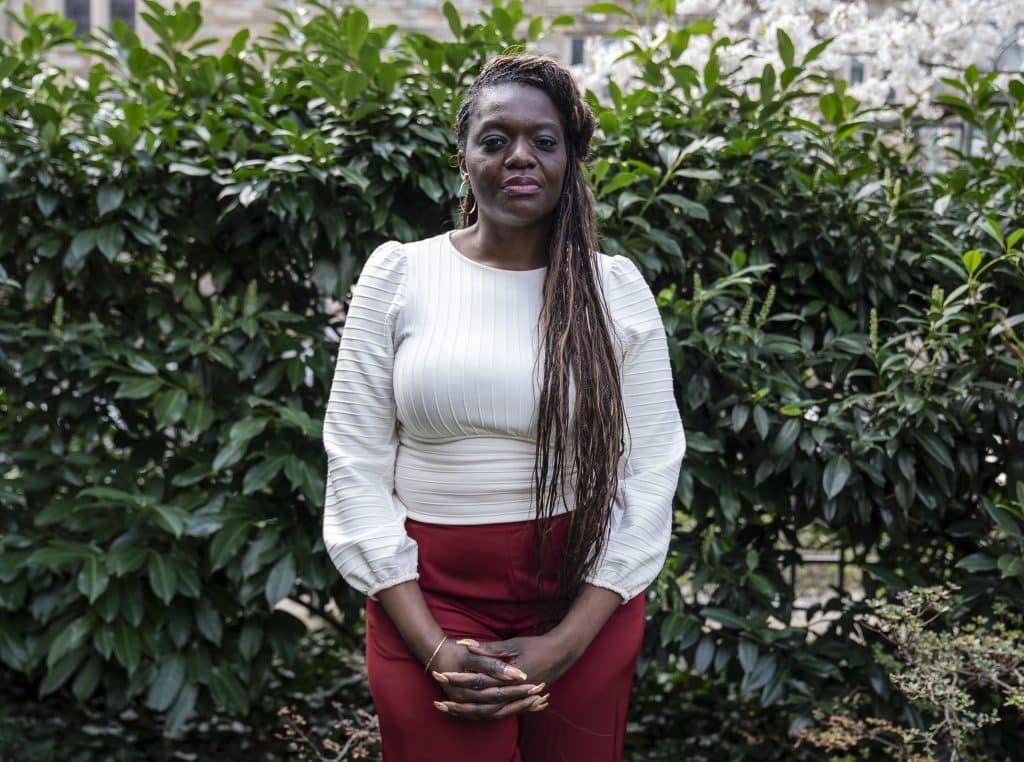 Alt text: A Black woman with long dark brown braids wearing a white blouse and red pants stands in front of a shrub with green leaves.