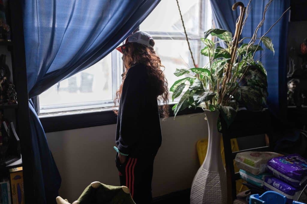 A woman with long, wavy red hair, whose face is not visible, looks out a window with blue curtains. Behind her is a tall vase with green leaves in it. A stack of diapers is on a chair.